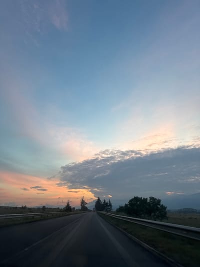 The image captures a serene twilight scene, likely either sunrise or sunset, over a road in Orizari, Bulgaria, viewed from what appears to be a moving vehicle. The expansive sky dominates the upper and left portions of the frame, showcasing a beautiful gradient of colors. On the far left and top, the sky is a clear, soft blue, gradually blending into lighter hues towards the center.

A significant band of dense, dark gray clouds stretches across the middle and right side of the sky, dramatically lit by the sun's warm glow. These clouds are edged with bright whites and oranges, suggesting the sun is either rising or setting behind them. Below and to the right of these darker clouds, the sky bursts with vibrant orange, pink, and yellow tones, indicative of the sun's low position. Further into the lighter blue areas, wispy, scattered clouds exhibit delicate pink and blue tinges.

The lower right quadrant of the image features a dark asphalt road, possibly a highway or rural main road, receding into the distance. A metallic guardrail runs along the right edge of the road, separating it from the roadside landscape. Alongside the road and extending into the middle ground, dark green trees and varied vegetation are visible, some appearing as silhouettes against the brighter sky. In the far distance, faint outlines of rolling hills or mountains are discernible beneath a hazy sky. The overall scene evokes a sense of tranquil travel through a picturesque landscape at a beautiful time of day. There is no visible text in the image.