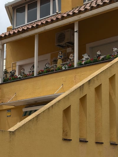 The image shows a building in Sant Andreu de Llavaneres, Spain. The structure has a beige-yellow exterior with a tiled roof and white window frames. The building's architectural details include recessed rectangular shapes in the facade. Along a balcony or ledge, figurines of people and gnomes are arranged amongst greenery and flowers. An air conditioning unit is mounted on an external wall. A sign is visible on a lower part of the building.