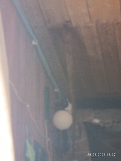 The image depicts an indoor scene, likely a dimly lit attic or storage space, with visible wooden beams and some electrical wiring. A round, white object, possibly a light bulb or a small sphere, is centrally located and appears to be out of focus. The overall lighting is low, with some light sources creating glare and shadows. The image also contains a timestamp in the bottom right corner: "26.06.2026 18:37", suggesting it was taken on June 26, 2026, at 6:37 PM. No people are visible in the frame, and the context of Novovolynsk, Ukraine, provides a geographical location without specific visual cues within the image itself. The scene appears still, with no discernible activity taking place. The quality of the image is grainy and somewhat blurry, making it difficult to discern finer details of the surroundings or objects.