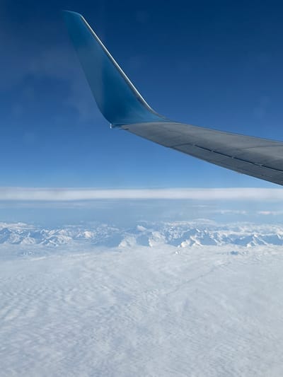 The image presents an aerial view from an airplane window. The photo is rotated 90 degrees clockwise, making the horizon appear vertical. On the left side of the frame, the aircraft's wing is prominently visible, extending from the top towards the lower center. The wing's light blue-grey underside and upward-curving wingtip are clearly discernible against the deep blue sky.

To the right of the wing, the expansive blue sky transitions into a rugged landscape of snow-capped mountains and dense clouds, which occupies the entire right half of the image. The mountain range features sharp, angular peaks and deep valleys, partially obscured by a blanket of white, textured clouds that extend further to the right. A faint, lighter band of haze or atmospheric layer runs vertically along the apparent horizon line.

The scene depicts bright, clear daytime conditions at high altitude. The provided context places this view over or near Novoye, Russia, suggesting a flight path over a mountainous region of Russia. There is no visible text in the image.
