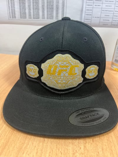 A black UFC championship belt-themed baseball cap with a flat brim is placed on a wooden surface. The cap features an embroidered gold and silver UFC championship belt design on the front. A circular black sticker with the Yupoong Snapback logo and "Yupoong Since 1974" is adhered to the underside of the brim, partially visible. In the background, a white document with columns of text and numbers is partially visible, along with a clear glass bottle with a yellow liquid and a white label. The scene appears to be indoors, possibly in an office or storage area. The city of Habshan, United Arab Emirates, is not visible in this image.