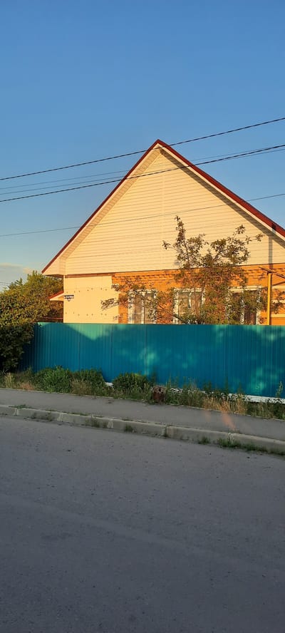 The image shows a side view of a two-story house with a light-colored, vinyl-sided upper story and a brick-patterned lower story. A dark red roof with white trim is visible. A bright turquoise metal fence runs along the front of the property, with green vegetation growing behind it. A tree with green leaves is positioned in front of the house, casting shadows on the fence. Power lines cross the sky above the house. The scene appears to be in a residential area, with a paved road and sidewalk in the foreground. The location is identified as being in the city of Shakhty City District, Russia.