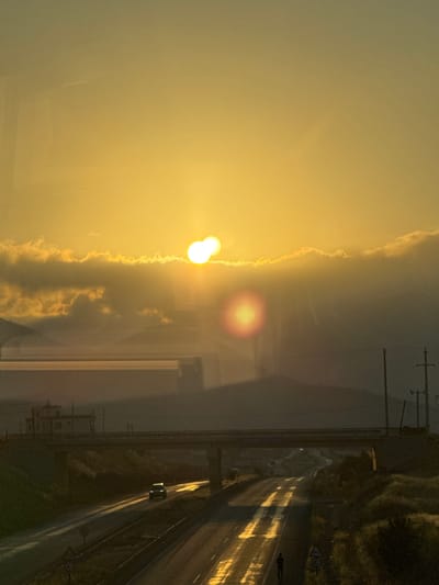 The image captures a hazy sunset over a highway in Silifke, Turkey. The sun, a bright orange orb partially obscured by clouds, casts a warm glow across the sky and reflects off the wet asphalt of the road. A multi-lane highway curves into the distance, with a bridge spanning over it. A dark-colored SUV is visible driving on the highway, its headlights cutting through the dim light. In the foreground, a person is standing on the side of the road, facing away from the camera. The landscape appears somewhat rugged with hills and vegetation visible on either side of the road. A few utility poles and wires are also present. The overall atmosphere is serene and somewhat melancholic, with the fading light creating a sense of stillness.