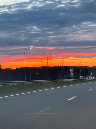 The media file shows a sunset scene along a highway. The sky is ablaze with vibrant hues of orange, red, and pink, transitioning to darker blues and grays as it extends towards the left of the frame. Wispy clouds are visible against the colorful sky. Along the edge of the highway, several lampposts with lit bulbs stand at intervals. A line of trees forms a dark silhouette against the sunset, separating the sky from the road. The highway itself is asphalt, with white lane markings visible. A few vehicles, appearing as headlights and taillights, are traveling on the road, suggesting it is either dusk or early evening. The overall atmosphere is serene and picturesque, capturing the beauty of a sunset over a travel route. The location context indicates this scene is in Bogorodskoe, Russia. No visible text is present in the image.