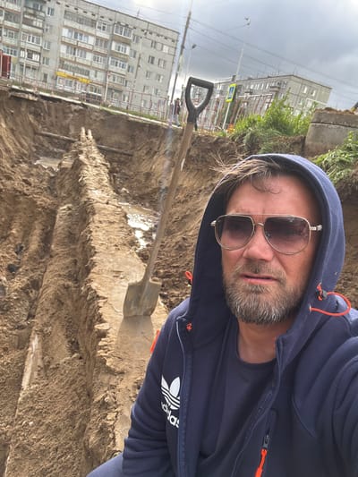 The image captures a man in the foreground, wearing a navy blue hooded jacket over a dark t-shirt, and aviator-style sunglasses. He has a beard and his hair is windblown. Behind him, a large excavation trench dominates the scene. The trench reveals layers of soil and what appears to be underground piping. A shovel stands upright in the trench, suggesting ongoing work. In the background, a multi-story apartment building and some fencing are visible, indicating an urban or suburban setting. The sky is overcast. The location is identified as Usinsk, Russia.