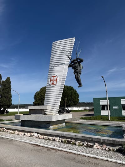 The image displays the Monument to the Paratrooper (Monumento ao Paraquedista) situated within a military compound in Praia do Ribatejo, Portugal, under clear daylight. The central subject is a multi-component monument resting on a rectangular, light-grey stone fountain structure. A tall, tapering, ribbed white vertical element, approximately 10-12 meters high, forms the primary support, featuring a circular emblem with a red Cross of Christ on a white background on its lower-left side. Suspended from the upper right of this vertical element by multiple thin metal rods, a dark bronze statue of a paratrooper is depicted in mid-descent, wearing a helmet, uniform, and backpack, with an upward gaze. The fountain basin beneath the monument contains water, revealing a colorful mosaic tile pattern on its floor. To the left of the monument are several tall, dark green coniferous trees. To the right, a dark green, single-story building with white window frames and a visible sign is present, alongside a tall, curved grey street lamp. A dark green military vehicle is partially obscured behind the monument's base. The foreground consists of a paved road with a cobblestone curb. The sky is clear blue with sparse, wispy white clouds. No human subjects are present in the immediate vicinity.
