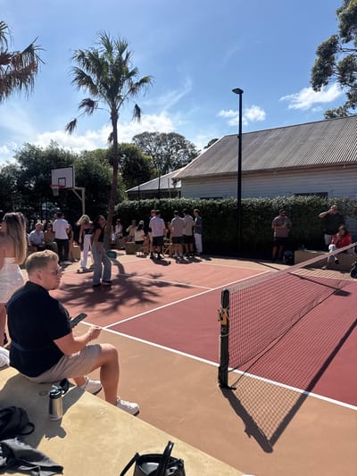 A sunny outdoor scene depicts a tennis court marked with red and tan sections, a net standing prominently. A group of people gathers near the court, some seated on chairs. A palm tree adds a tropical touch, contrasting with the structure of a building visible in the background. A basketball hoop is also present, indicating a multi-purpose sports area. Someone is sitting near the edge of the tennis court.