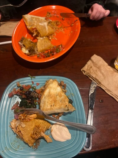 The image shows a table in a restaurant with two plates of food on it. The first plate is orange and contains two folded quesadillas, one of which has been partially eaten. The second plate is blue and contains a larger quesadilla, black beans, spinach, tomatoes, and a small dish of white sauce. A silver fork and a silver knife are on the table. A brown paper napkin is on the table to the right of the plates. The coordinates (latitude: 41.021, longitude: -73.635) correspond to a location in Greenwich, Connecticut, United States, but the image does not provide any information about the restaurant's location.