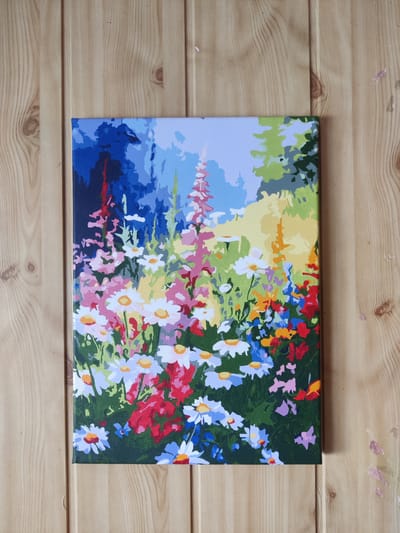 This is a top-down, medium shot of a paint-by-numbers canvas depicting a colorful flower garden. The canvas is placed on a wooden surface, likely a table, with visible wood grain and knots. The painting features a variety of flowers in shades of red, pink, white, blue, and yellow, with green foliage. In the background, there are stylized trees and a blue sky. The paint application is clearly visible, with distinct blocks of color indicating the paint-by-numbers format. No people are present in the image. The wooden surface suggests an indoor setting, and there are no elements in the image that would indicate a specific location within Koziatyn, Ukraine.