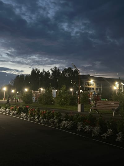 The image captures a nighttime scene in a park or recreational area in Bogotol, Russia. The sky is overcast with dark, textured clouds, suggesting dusk or night. Streetlights illuminate the area, casting a warm glow on the paved walkway and adjacent flowerbeds. A row of red and white flowers lines the walkway, with lush green bushes behind them.
In the background, a multi-story building with visible windows and lights stands under the evening sky, possibly a residential or institutional building. Adjacent to the building is a playground with visible structures, suggesting it's a family-friendly area. There are streetlights scattered throughout the scene, some on tall poles and others integrated into the playground area.
A wooden bench is placed in the foreground, offering a spot for rest. Two individuals are faintly visible in the middle ground; one is walking on the paved path, and another appears to be interacting with playground equipment. The overall atmosphere is calm and serene, illuminated by artificial light against the dark sky.