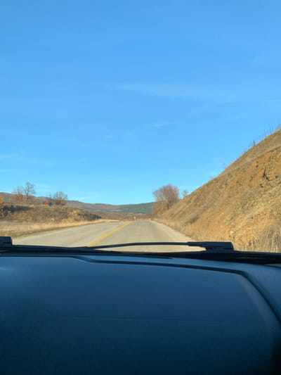 The image presents a driver's-eye view from inside a vehicle, looking out onto a road winding through a dry, hilly landscape in Grevena, Greece. The perspective is through the car's windshield, with the dark dashboard and a segment of a windshield wiper visible on the right side of the frame.

A paved road, marked by a solid yellow double line down its center, curves gently into the distance, suggesting a path through mountainous or rural terrain. On both sides of the road, the land rises into steep, dry hills. These hills are covered with sparse, light brown vegetation and scattered, bare deciduous trees, indicating a dry season, possibly late autumn or winter. The overall color palette of the landscape is dominated by earthy tones of brown, yellow, and orange.

Above the rugged terrain, the sky is a bright, clear blue, stretching across the upper left quadrant of the image. A few faint, wispy cloud formations or contrails are visible, but the sky is predominantly clear and unblemished, suggesting sunny weather conditions. The bright lighting indicates it is daytime, likely morning or early afternoon.

No individuals are visible in the scene, conveying a sense of serene solitude on the road. There is no discernible text within the image. The photograph captures a tranquil moment of travel through a natural, rugged environment.
