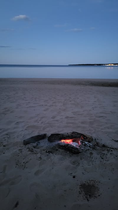 This is a photograph of a beach with a campfire burning on the sand. The scene is set at dusk, with the sky a deep blue and the water a calm, reflective surface. The campfire is in the foreground, with logs smoldering and glowing embers. The sand is a light tan color, with footprints visible in some areas. In the distance, across the water, there is a dimly lit shoreline with lights from buildings. The overall impression is one of a peaceful and serene evening by the water.