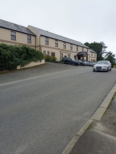 The image captures a frontal view of the Castles of Oir Hotel in Annagry West, Ireland, on an overcast day. The hotel is a two-story building with a beige facade, a dark grey tiled roof, and numerous brown-framed windows. A prominent sign above the entrance reads "CASTLES OF OIR HOTEL". Green hedges line the front of the hotel, partially obscuring the lower level. In front of the hotel, parked on a sloped asphalt area, are several cars: a dark grey SUV on the left, a black sedan behind it, and a silver Audi A3 in the foreground, which is in motion, approaching the viewer. The road in front of the hotel is also asphalt and slopes downwards. The sky is a uniform grey, indicating cloudy weather. A concrete curb and gutter are visible along the right side of the road.