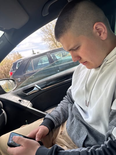 A young man with short hair is seated in a car. He is wearing a grey and white hoodie. He is holding a smartphone in his hands. Outside the car, another vehicle is visible. The location is impossible to discern from the image.