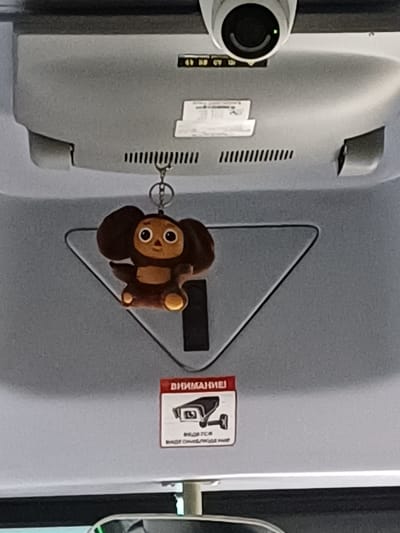 A plush toy keychain of a cartoon character, Cheburashka, is hanging from a metal ring attached to the ceiling of a vehicle. The toy is brown with large ears and eyes. Below the toy, a red and white sign with an image of a security camera and Russian text is affixed to the ceiling. The text on the sign reads "ВНИМАНИЕ!" which translates to "ATTENTION!". The ceiling of the vehicle appears to be made of grey plastic, and a security camera is visible at the top of the image, positioned above the toy and the sign. The background shows the interior of the vehicle, suggesting it could be a bus or a van. The image is taken from a low angle looking upwards.