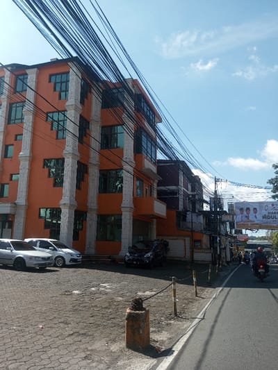 The image presents a street view in Ban Phonhxavanh, Laos, under bright daylight. A prominent multi-story building, likely five floors, painted a vivid orange, occupies the left and central portion of the frame. Its facade is distinguished by multiple vertical white pilasters or columns with decorative capitals, interspersed with numerous large, dark-tinted rectangular windows.

In front of the orange building, a paved or cobblestone parking area holds three parked vehicles: a silver sedan, a white hatchback, and a black SUV or minivan closer to the building's entrance. Overhead, a dense network of black utility wires crisscrosses diagonally across the sky and partially obscures the building.

Along the edge of the parking area, a short, weathered concrete bollard with an attached metal chain serves as a barrier, connected to a similar wooden post further down. To the right, an asphalt road extends into the distance, with a white line marking its edge. A motorcyclist wearing a red helmet and dark clothing is visible in the right lane, heading away from the viewer. Other vehicles, primarily motorcycles, are discernible further down the road.

Beyond the orange building and along the road, smaller, darker-colored structures are visible. A large billboard mounted above the road displays multiple individuals and text, notably "SELAMAT HARI RAYA IDUL FITRI". The sky is clear blue with sparse white clouds, indicating a sunny day.