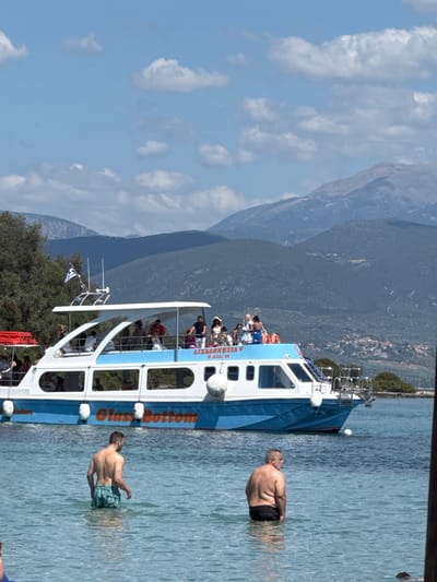 In Lichada, Greece, a "Glass Bottom" boat is docked in clear, shallow blue water. The boat is white and blue with several passengers visible on its upper deck. Trees and mountains form the backdrop. In the foreground, two men stand in the water. One man, facing away from the camera, wears patterned swim trunks. The other man, also facing away, wears black swim trunks. The water is calm and transparent.