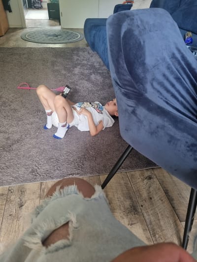 A young child is lying on a grey carpet, underneath a large, dark blue, plush chair. The child is wearing a white t-shirt with a cartoon character on it, white shorts, and white socks with blue accents. They are holding a pink toy with a comb-like attachment and a black object with white buttons, possibly a remote control. The child appears to be relaxed or asleep, with their head resting on their arm and their legs bent.

The background shows a wooden floor and a portion of a light blue fuzzy rug. To the left, there is a glimpse of kitchen cabinetry and a round rug with a star pattern. The lighting suggests it is daytime, and there are no indications of specific weather conditions. The overall scene appears to be an indoor living space, possibly a living room or playroom. No visible text is present in the image.
