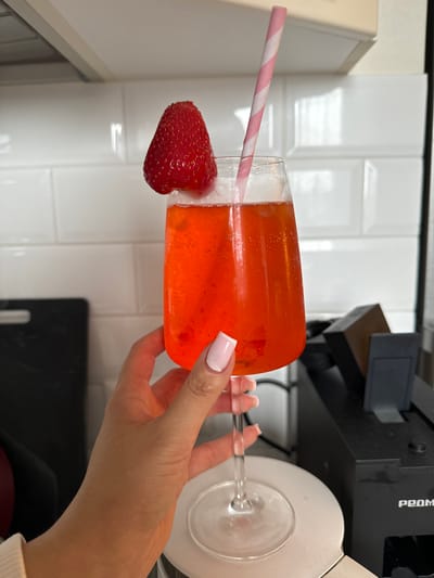 The image shows a hand holding a glass filled with an orange liquid, likely a cocktail. A large strawberry is attached to the rim of the glass, and a pink striped straw is visible inside the glass. The background includes white subway tiles, a portion of a coffee machine, and the edge of a white cabinet. The scene gives no indication of the location in Gribovo, Russia.
