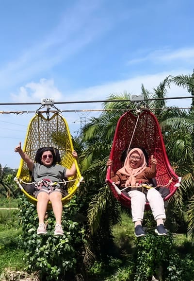 In Bintang Meriah, Indonesia, two women are seated in suspended, colorful hanging chairs, enjoying an outdoor adventure. The woman on the left, wearing sunglasses and a grey top with a patterned skirt, gives a thumbs-up gesture. The woman on the right, wearing a light pink hijab and a brown top with white pants, also gives a thumbs-up. Both are secured by safety harnesses. The chairs are suspended from a cable line, suggesting they are part of a zip line or similar attraction. The background features lush green foliage, including palm trees, and a bright blue sky with some clouds. The scene suggests a recreational activity in a tropical or subtropical environment.