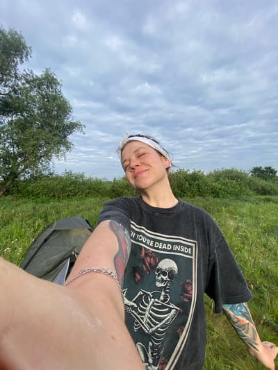 A young woman is taking a selfie outdoors in a grassy field with a cloudy sky. She is wearing a dark t-shirt with a skeleton graphic and the text "YOU'RE DEAD INSIDE" printed on it. She has a white bandana tied around her head and visible tattoos on her arms. To her left, a small green tent is pitched in the grass. In the background, there are trees and dense green bushes. The location is identified as the village of Abzanovo, Russia.