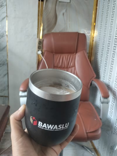 A hand holds a black tumbler with a logo that reads "BAWASLU BADAN PENGAWAS PEMILU UMUM" in front of a brown leather office chair. The tumbler is filled with a light-colored beverage, possibly coffee, with foam on top. The background features a wall with a repeating pattern and what appears to be a gold-trimmed column. The scene is indoors and is likely in an office setting in Rantopanjang, Indonesia, given the presence of the BAWASLU logo, which is the Indonesian Election Supervisory Agency.