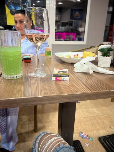 This image shows a close-up view of a wooden table in what appears to be a restaurant or bar. On the table are a glass of green liquid, a wine glass with rosé wine, a pack of cigarettes, a crumpled white napkin, a small plant in a white pot, and a plate with some food remnants. A woman in a blue and white striped shirt is partially visible in the background, to the left of the frame. The floor appears to be tiled. The overall impression is of a casual dining setting, possibly in Zlataritsa, Bulgaria, given the context provided, though the location is not explicitly depicted in the image itself.
