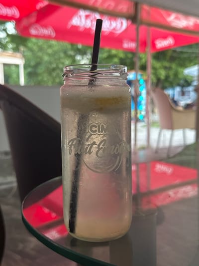 A chilled glass jar with a straw and a yellow liquid is placed on a round glass table. The jar has a logo that reads "CIM Fruit Emotion." In the background, there are red umbrellas, green foliage, and a blurred outdoor setting that suggests a cafe or restaurant. The lighting is natural, indicating daytime. The overall scene is focused on the drink and its immediate surroundings, with the background out of focus. The location is identified as the city of Kubrat, Bulgaria.
