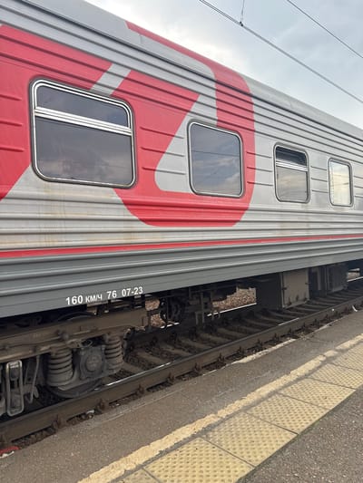 The image shows a side view of a Russian Railways train car at a station. The train car is primarily gray with large red markings. There are several windows on the side of the train car, reflecting the overcast sky. Below the windows, the car is marked with "160 KM/H 76 07-23" in white lettering. The train is on a set of tracks, with ballast and ties visible. A yellow tactile paving strip is present along the edge of the platform, which is made of asphalt and concrete. The scene appears to be a railway station in Mariinsk, Russia. There are no people visible in the image.
