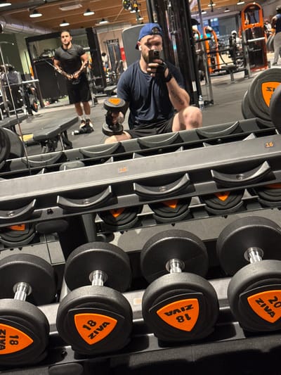 A person takes a mirror selfie in a gym setting. The individual is wearing a blue cap, a dark t-shirt, and workout gloves. They are holding a dumbbell in one hand. The background shows gym equipment, including a rack filled with ZIVA-branded dumbbells, ranging from 16 to 20. The gym appears to be well-equipped and modern. The photo is taken at an angle, offering a partial view of the gym's interior.