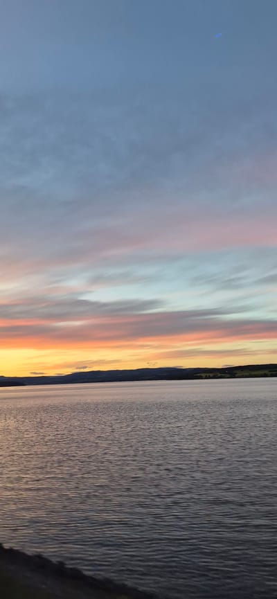 This image captures a serene sunset over Mjøsa, Norway's largest lake, with the town of Gjøvik visible in the distance on the far shore. The sky is painted with a gradient of soft colors, transitioning from a pale blue at the top to hues of pink, orange, and yellow closer to the horizon. Wispy clouds stretch across the sky, catching the last rays of sunlight. The water of Mjøsa is calm, with gentle ripples reflecting the colors of the sky. In the background, a dark, silhouetted mountain range forms the horizon, punctuated by a few scattered lights from the town of Gjøvik. The foreground shows a dark, blurred shoreline. The overall impression is one of tranquility and natural beauty.