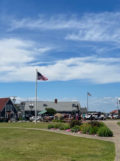 This is a daytime, eye-level, outdoor, vertical shot taken in Oak Bluffs, Massachusetts, United States of America. In the foreground, a large expanse of green grass is visible, with a paved pathway curving through it. A flowerbed with pink and yellow flowers is situated near the pathway. In the midground, an American flag waves from a tall flagpole. Behind the flag, several buildings with gray roofs are visible. One building in the background has a prominent red roof and dark siding. A parking lot with several vehicles, including white SUVs and a Jeep, is located behind the buildings. A group of people are gathered near picnic tables and a large boulder in front of the buildings. Another smaller American flag is visible on a pole in the background, to the right of the main flagpole. The sky is blue with scattered white clouds. The overall impression is a bright, sunny day in a coastal town.
