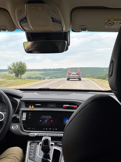 The image shows the interior of a car driving on a road, with a landscape visible through the windshield. In the foreground, part of the dashboard and seats are visible. On the dashboard, there is a digital display screen showing music information and a QR code. Two yellow duck figures are placed on the dashboard near the air vents. The steering wheel is partially visible on the left.
In the middle ground, a dark-colored SUV is driving ahead on the same road. The road itself appears to be asphalt and is flanked by grassy shoulders. The background features rolling green hills covered with trees, indicating a rural or semi-rural environment. The sky is overcast with some patches of blue. The overall lighting suggests daytime.
The image is taken from the perspective of a passenger or driver inside the car. The text on the dashboard display appears to be in Russian, with the song title "Тёмный учитель" (Dark Teacher) and artist "Король и Шут-Театр демона" (Korol i Shut - Demon Theater) visible. The temperature is displayed as 22.0°C.
The location is stated to be Дюсьметьево, Russia. This context suggests the scene is taking place on a road within or near this locality.