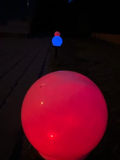 An outdoor, low-light photograph shows a series of glowing orbs lining a path. The closest orb, in the foreground, is large and bright red. Further back, a smaller blue orb is visible, topped by a tiny red orb. In the distance, another red orb is faintly visible. The background is dark, with some indistinct structures that might be buildings or fences, suggesting an urban or suburban setting. The presence of glowing orbs suggests decorative lighting, possibly for an event or holiday. This scene was captured in Rumia, Poland.