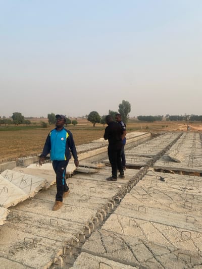 Three men stand on concrete slabs. The person in the foreground wears a blue and black track suit. Two other men are behind him, one in black. The slabs appear to be part of some infrastructure construction, possibly for erosion control. The background shows a flat, arid landscape with sparse vegetation and a hazy sky.