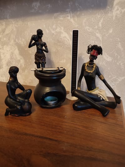 In this media file, three decorative figurines depicting people are arranged on a wooden surface. The figurines are black and have an African aesthetic. The first figurine, on the left, is a woman kneeling and holding a pot. The second figurine, in the center background, is a woman standing and carrying a baby on her shoulder. The third figurine, on the right, is a woman sitting cross-legged. She wears a red headband and gold jewelry.

In front of the standing figurine, there is a black ceramic oil burner. Inside the burner, a small green candle is lit. A thin wooden stick rests across the top of the burner.

Behind the figurines, a textured wallpaper is visible. A thin, black and white striped object, possibly a ruler or a bookmark, stands upright between the standing figurine and the oil burner.

The overall scene appears to be a still life arrangement on a shelf or tabletop. There are no overt indications of time of day or weather, nor any strong emotional cues. The location is identified as Abinsk, Russia. No visible text is present.