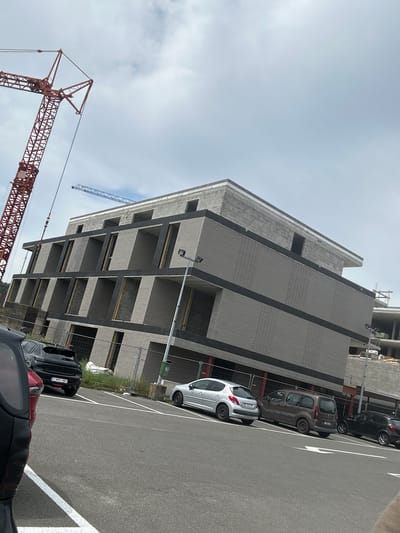 The image depicts a construction site in Herstal, Belgium, under a cloudy sky. A large, multi-story building, still under construction, dominates the scene. Its facade features light-colored brickwork interspersed with dark horizontal bands. Several levels are visible, with open, unfinished balconies or window openings.
In the foreground, a red construction crane is prominently displayed, its intricate metal lattice structure reaching upwards. Another, smaller blue crane is visible in the background, to the right of the building.
Parked in front of the building are several vehicles, including a silver hatchback, a brown van, and a dark-colored car. The parking lot is paved and marked with white lines. Some overgrown grass and greenery are visible near the base of the building, adjacent to the parking area. Fencing encloses parts of the construction site.
The time of day appears to be daytime, given the ambient light, and the weather is overcast, suggesting a cloudy or slightly hazy day. No people are clearly visible in the image, but the presence of vehicles and construction equipment indicates ongoing activity at the site. Visible text includes license plates on the cars, such as "1-NV-881" on the rear of the dark car.