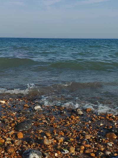 The image depicts a serene coastal scene at Worthing, United Kingdom. The foreground is dominated by a pebbled beach, with a variety of stones in shades of brown, grey, white, and orange, some wet from the incoming tide. Small patches of green seaweed are scattered amongst the pebbles.

The middle ground features the gentle, rippling waves of the sea, with white foam cresting as they approach the shore. The water is a deep blue-green, reflecting the clear sky.

In the distance, the horizon is a sharp line separating the sea from the sky. The sky is a pale blue with subtle hints of clouds. A faint, distant line of what appear to be wind turbines is visible on the horizon, a common sight in the coastal areas of the UK.

The time of day appears to be late afternoon or early evening, suggested by the soft, clear light and the calm nature of the sea. The weather is clear and pleasant, with no strong winds or rain indicated.

The overall mood conveyed by the image is one of tranquility and natural beauty. There are no people or specific man-made objects in the frame, focusing the viewer's attention entirely on the natural elements of the beach and the sea. There is no visible text in the image.