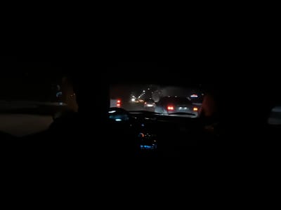 The image shows the interior of a car at night. The dashboard is visible, including the steering wheel and gauges. Outside the car, other vehicles are on the road, their headlights and taillights illuminated. The scene suggests that the car is in traffic at night. The overall tone is dark.