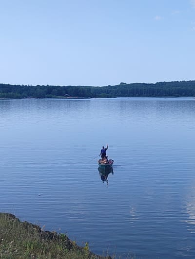 An image taken from a slightly elevated, foreshore perspective shows a calm, large body of water, likely a lake, with a distant, treed shoreline. In the middle ground of the water, a small, green boat is occupied by several individuals. One person stands at the stern, facing the viewer and waving, while at least two others are seated within the boat. The water surface displays ripples, indicating a gentle breeze. The sky above is clear and blue. The foreground shows a rocky and grassy embankment. The scene appears to be a recreational setting, possibly in Roxton Pond, Canada, given the context.