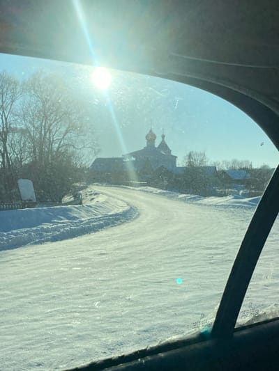 This image depicts a snow-covered rural landscape in Пліскі сельскі Савет, Belarus, captured from the interior of a vehicle through a streaked window. A winding road, covered in white snow and flanked by snowbanks, recedes into the mid-ground. An Eastern Orthodox church, characterized by a dark structure and two prominent golden onion-shaped domes, is centrally located in the mid-ground, surrounded by smaller, dark-roofed buildings typical of a village. Bare deciduous trees, lightly covered in snow, line the left side of the road and extend into the background. The sky is clear and pale blue, with a bright sun positioned in the upper left quadrant, generating substantial lens flare across the upper portion of the scene. Dark vehicle interior elements, including the headliner and window frame, are visible along the top and right edges of the frame. A small, circular cyan light artifact is present on the lower right section of the window.