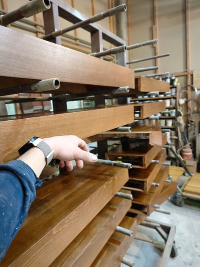 The image depicts an indoor scene within a woodworking workshop or factory located in Negorci, North Macedonia. In the foreground, a light-skinned left hand, wearing a dark blue long-sleeved garment with a slightly frayed cuff and a smartwatch with a black body and light gray fabric strap, extends towards a finished wooden plank. The hand's position suggests interaction or inspection of the plank.

The central focus of the image is a multi-tiered drying rack system constructed from dark brown frames and numerous horizontal metal tubes. Stacked horizontally across these tubes are multiple rectangular wooden planks or beams, all uniformly finished with a glossy, medium-brown stain or varnish, showcasing visible wood grain. These planks appear to be in a post-finishing drying stage.

The background, out of focus, reveals additional workshop elements. These include shelving units stocked with various containers, likely paints, solvents, or supplies. Further back, equipment such as a wheeled cart, an upright object resembling a red fire extinguisher, and more stacked wood products or components are partially visible, indicating an active industrial environment. The walls are light-colored, possibly with exposed vertical wooden studs.