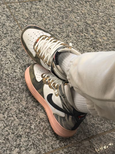 The image shows a close-up, overhead shot of a person's legs and feet. The person is wearing white sweatpants and white socks. Their feet are shod in Nike Air Force 1 shoes. The shoes have a white vamp, camo print on the quarters and around the toe, black swooshes, and salmon-colored soles. The setting is a gray terrazzo floor. The location is not indicated to be in the city of Pyrzowice, Poland.