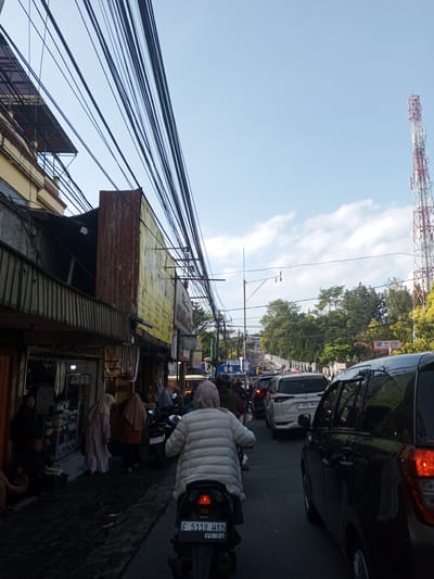 The image depicts a bustling street scene, likely in an urban area. A densely packed assortment of overhead power lines crisscross the sky above a line of shops and buildings on the left side of the frame. People are seen walking and congregating on the sidewalk in front of the shops.

In the foreground, a person is riding a scooter with license plate F 5118 HAB down the street. A line of cars and other vehicles is visible on the right, indicating traffic congestion. In the distance, a cell phone tower rises above the trees, suggesting the presence of communication infrastructure.

The sky is partly cloudy, and the overall lighting suggests it is daytime. It is impossible to confirm this image is of Nhot Chang, Laos.