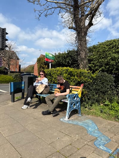 A daytime street scene in Kenilworth, United Kingdom, depicts two individuals seated on a public bench. The location is identifiable by a visible "Boothroyd LET" sign for a real estate agency, suggesting proximity to Boothroyd & Co, 18 The Square, Kenilworth.
The scene features a paved concrete sidewalk beside a road, with a dense green hedge and a bare-branched tree forming the background. Bright sunlight casts distinct shadows.
On the left side of a public bench, a female individual is seated. She wears dark sunglasses, a light blue long-sleeved top, dark trousers, and white athletic shoes. Her head is tilted downwards, focused on a white smartphone held in both hands. A light-colored canvas-style bag rests on the bench beside her.
To her right, a male individual is seated on the same bench. He wears a black t-shirt with a graphic design, olive green cargo pants, and black strapped sandals or open-toed shoes. He holds a light brown cylindrical object in his hands, looking down at it.
The bench possesses ornate blue-painted metal ends and wooden slats painted in multiple colors, including blue, green, yellow, orange, and red. The paint on the metalwork shows signs of wear and chipping. On the concrete sidewalk directly in front of the bench, particularly extending from the right side, there is a light blue, wave-like chalk or painted art design.
In the background, to the left, a black traffic light pole is partially visible. A blue and black utility box is also present on the sidewalk closer to the road. A red brick building is partially obscured by the signage and tree.
