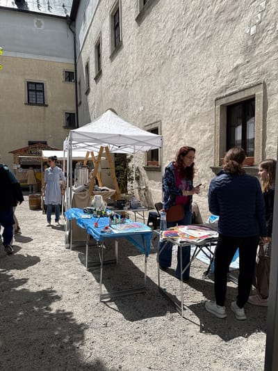 A daytime outdoor scene shows a small market or fair in the city of Častá, Slovakia, on a gravel surface. A woman with long red hair sits at a table displaying art, engaging in conversation with two standing individuals. Various products and items for sale are arranged on tables under a white canopy. A person in a white coat is standing by another table. Buildings surround the scene. Shadows cast by trees are visible on the ground. Other individuals are also present and appear to be browsing the market.