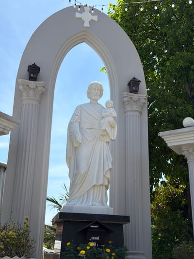 The image shows a white marble statue of Saint Joseph holding the child Jesus, positioned within a large, arched entryway. The statue is centrally located, with fluted columns flanking the arch on either side. Decorative lanterns are mounted on top of the columns. Above the arch, a white cross with four equal arms is visible against a clear blue sky. Green trees and foliage are present in the background. At the base of the statue, a dark rectangular pedestal is visible, with some yellow flowers in the foreground. A sign on the pedestal reads "XIN KHAI MỞ NHÀ", which translates to "Please open the house". The scene appears to be outdoors, likely in a religious or historical site in Hội Tín, Vietnam.