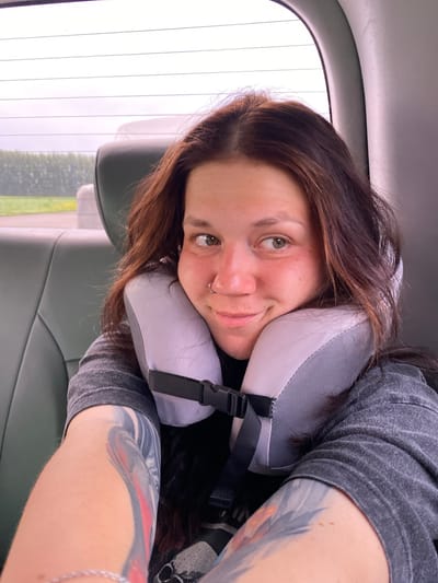 A woman with dark brown hair and fair skin is seen from the chest up, leaning against a grey neck pillow. She is wearing a dark grey t-shirt. Her left arm, with colorful tattoos, is visible in the foreground. She has a nose ring and is looking to her right with a slight smile. She is in the back seat of a vehicle, with the headrest of the seat in front of her visible. The background shows a window with a blurry view of trees and a cloudy sky. The image appears to have been taken on a road trip.