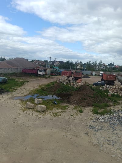 The image shows a construction or industrial site in Яльгелево, Russia. In the background, there are residential buildings and a large pile of gravel or sand. Several trucks, some with red dump beds, are parked in the area. A blue car is also visible. In the foreground, there's a dirt area with puddles, piles of dirt, grass, and scattered concrete blocks and rocks. The sky is cloudy.