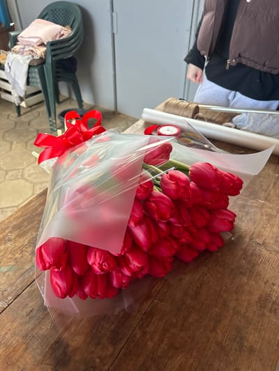 A large bouquet of approximately 50-60 vibrant red tulips, wrapped in a translucent plastic film over a white paper liner and adorned with a red satin bow, rests on a distressed wooden table. The scene is located in Trudobelikovskiy, Russia. In the upper right background, a person, visible from the torso down, wears a brown puffer vest over a dark shirt and light grey sweatpants, standing near floristry supplies. These supplies include a roll of brown twine, a roll of white wrapping paper, and a spool of red ribbon. To the left background, a stack of dark green plastic chairs holds light-colored fabric items. The wall behind is light grey, and the floor is a light-colored, possibly tiled or concrete surface. The setting appears to be an indoor workspace or flower preparation area.